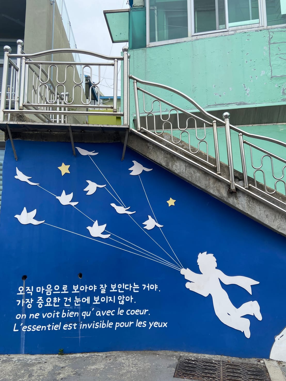 A Little Prince mural on a stairwell wall, showing his silhouette holding strings tied to a flock of birds, with a quote in Korean and French