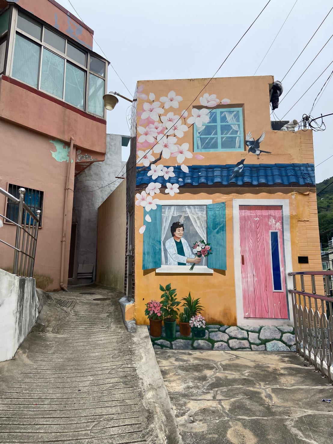 A mural of a woman holding flowers at a window, painted on a house wall with cherry blossoms above