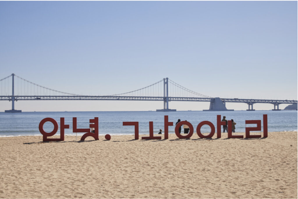 A large red 'Hello, Gwangalli' sculpture on the sand at Gwangalli Beach, with Gwangan Bridge in the background