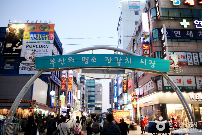 The arched entrance sign to Jagalchi Market in Busan, lit up at dusk with crowds passing underneath