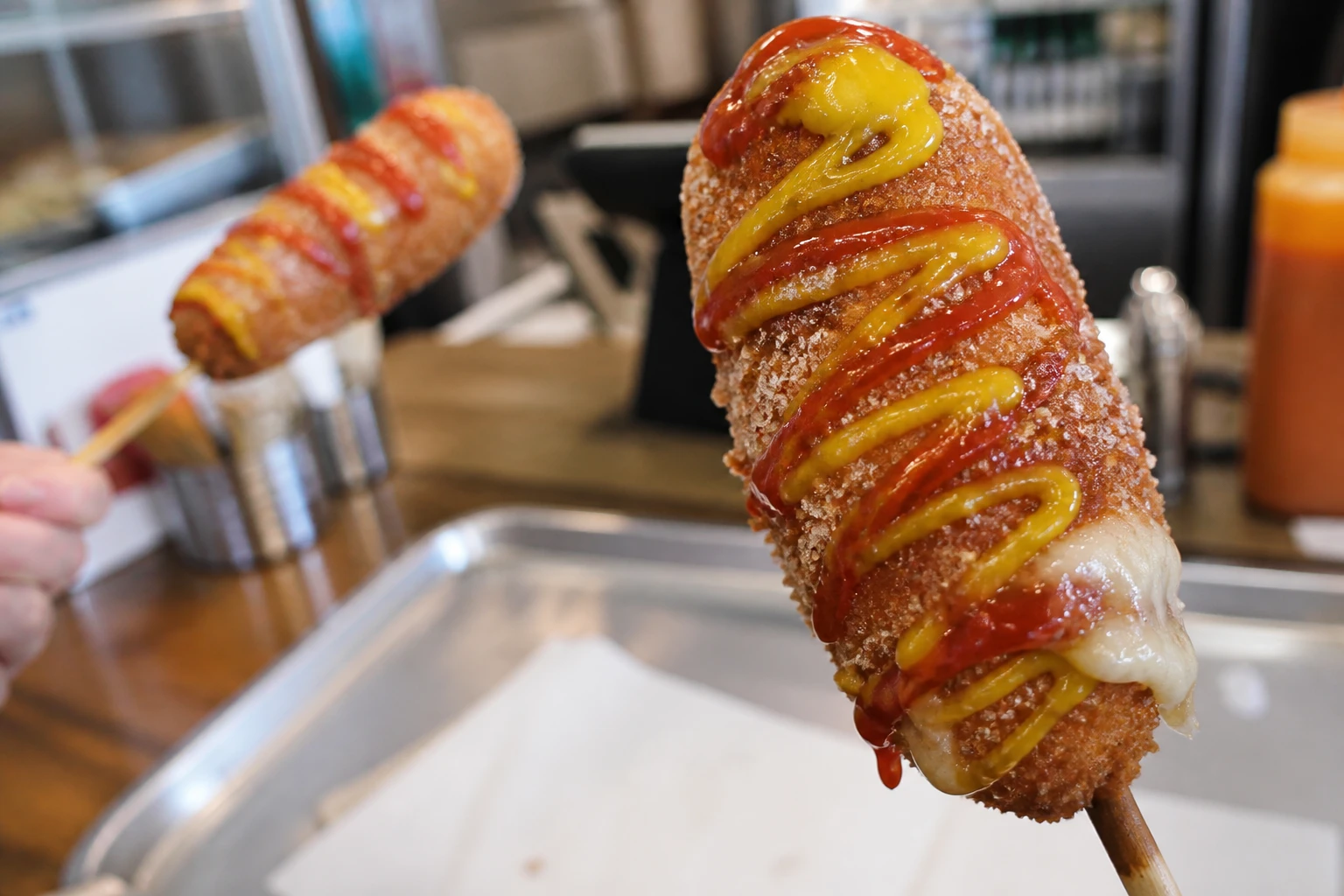 A sugar-dusted Korean corn dog drizzled with ketchup and mustard, cheese pull visible at the bottom, with a second corn dog in the background