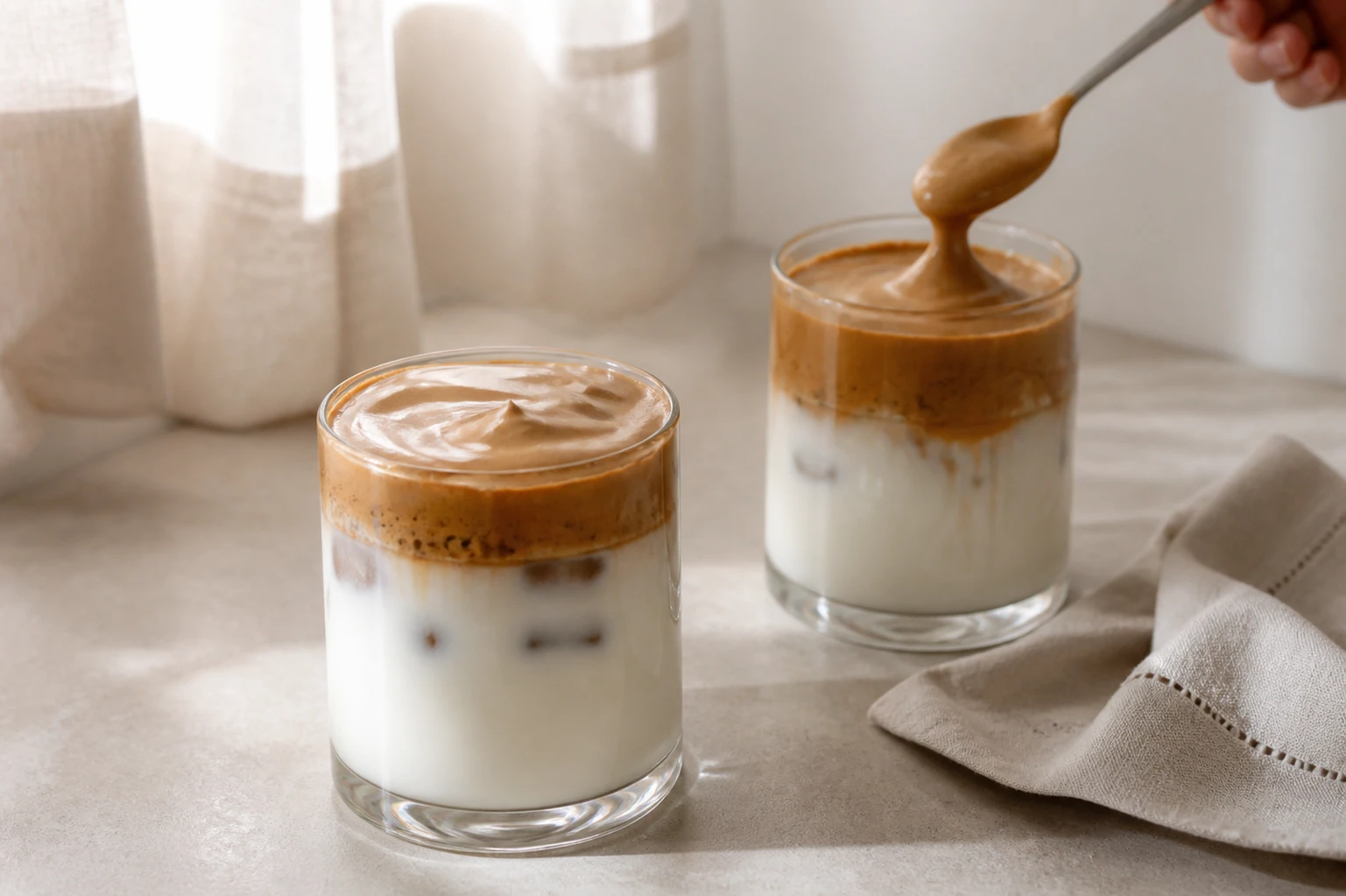 Two glasses of dalgona coffee with whipped coffee foam layered over milk, one being topped with a fresh spoonful of the foam