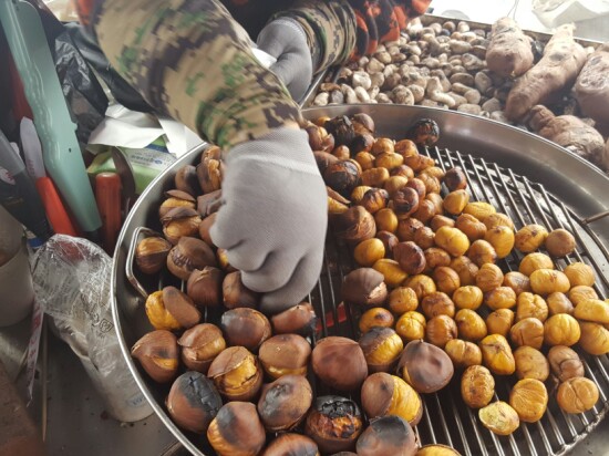 A vendor's gloved hand tending a pan of roasted chestnuts over hot coals at a Korean street stall