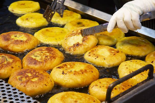 Rows of hotteok frying on a flat-top griddle, a gloved hand pressing one flat with tongs