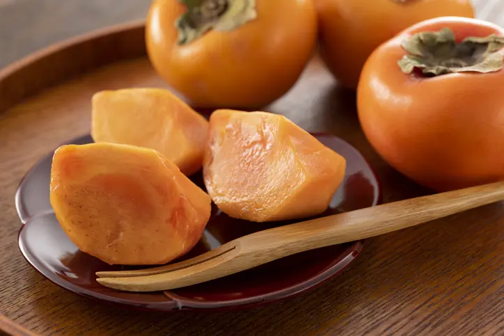 Whole ripe persimmons and a few cut open on a wooden tray, showing the soft orange flesh inside