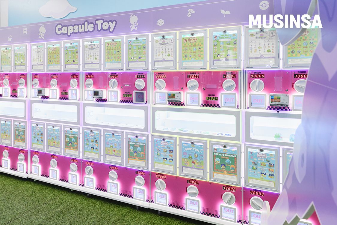 A long wall of pink and white Pokopia capsule toy machines with product posters for grip toks, acrylic dioramas, shaker keyrings and pin badges