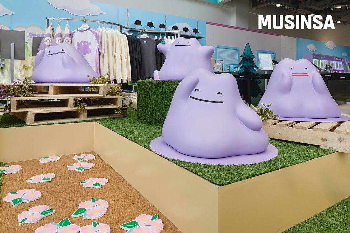 Four giant purple Ditto sculptures sitting on fake grass and wooden pallets in front of racks of Pokopia T-shirts and hoodies