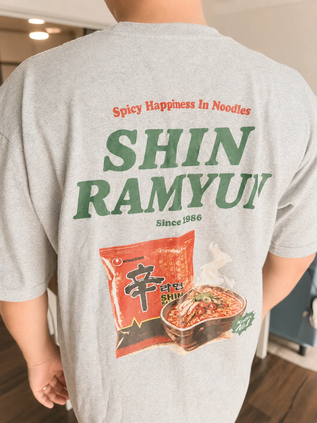 The back of the grey Shin Ramyun T-shirt, printed with Spicy Happiness In Noodles, the Shin Ramyun wordmark, and Since 1986
