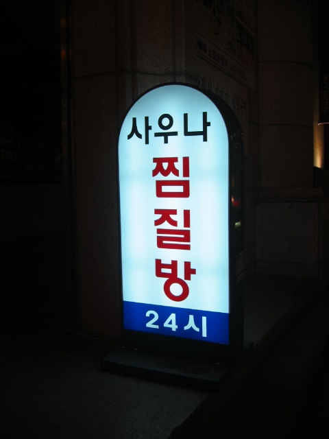 Lit-up street sign reading 사우나 찜질방 24시, sauna jjimjilbang open 24 hours, at night