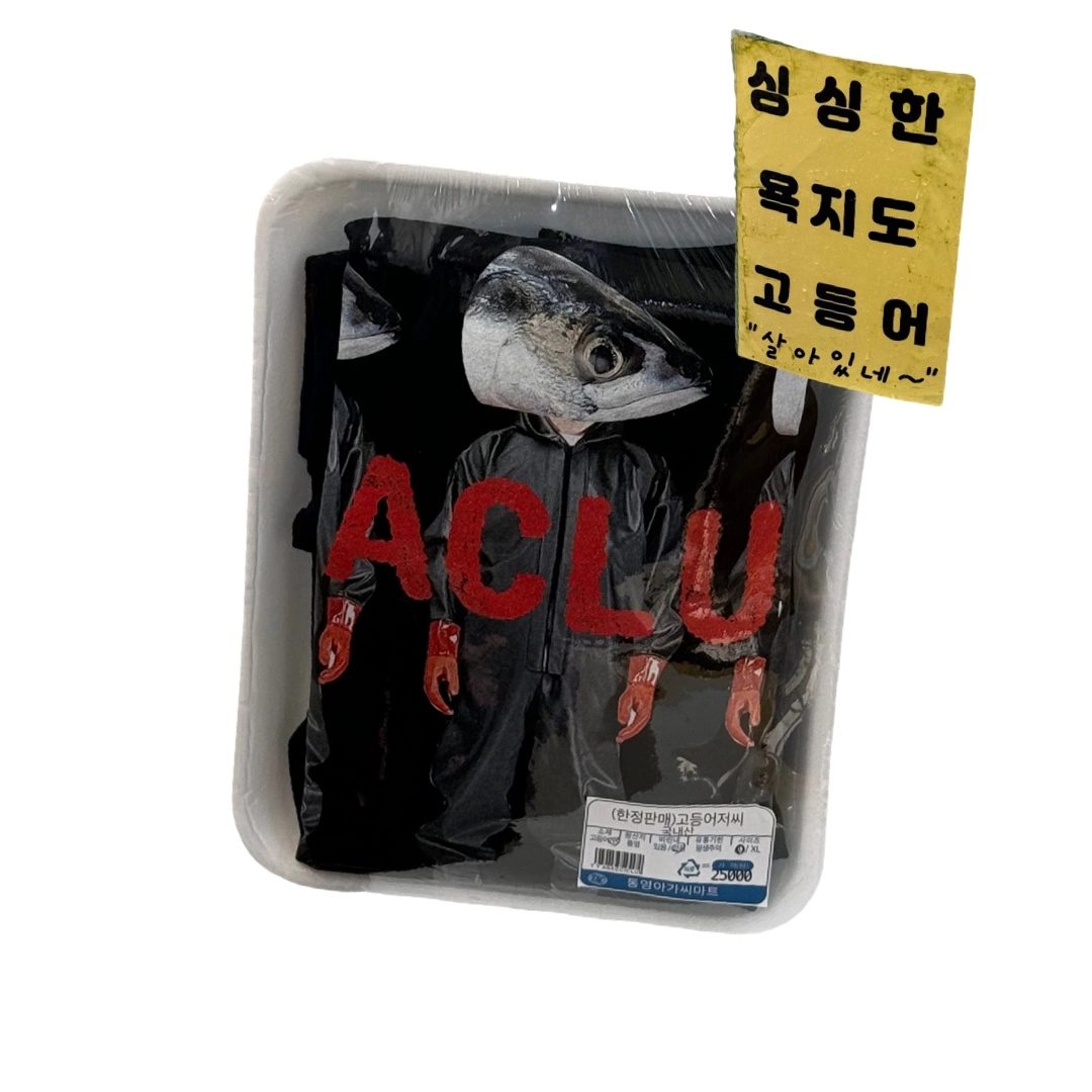 A black T-shirt with a mackerel print, shrink-wrapped on a foam tray with a yellow sign reading 싱싱한 욕지도 고등어